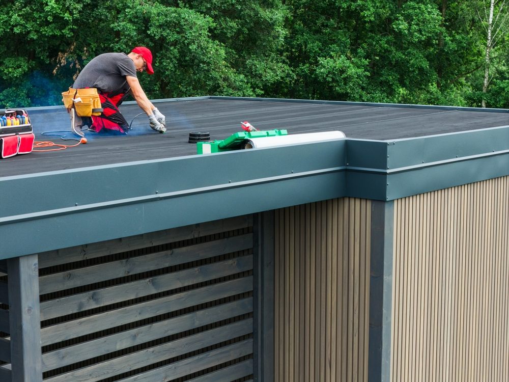 Flat Roofing Services In Ottawa. On a bright day, a worker is applying a protective coating to the flat roof of a contemporary building surrounded by lush greenery.